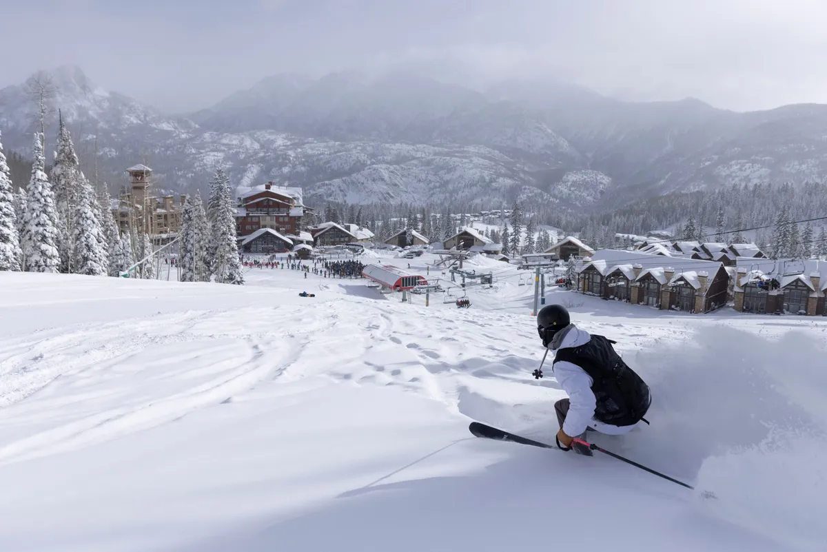 A dynamic scene showing a skier speeding down the snow-covered slopes with the resort buildings and pine trees in the background. The mountain landscape is prominent, and the skier’s movement conveys excitement and direct access to the slopes from the property.