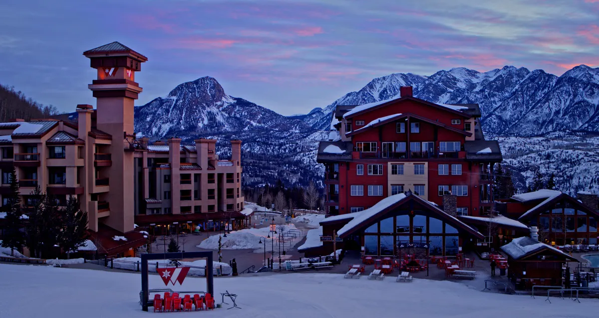 This wide-angle exterior image showcases a snow-capped mountain range with resort buildings nestled below. The landscape captures the serene beauty of the area, hinting at the proximity to winter sports and scenic relaxation options such as fireside gatherings after a day of adventure.