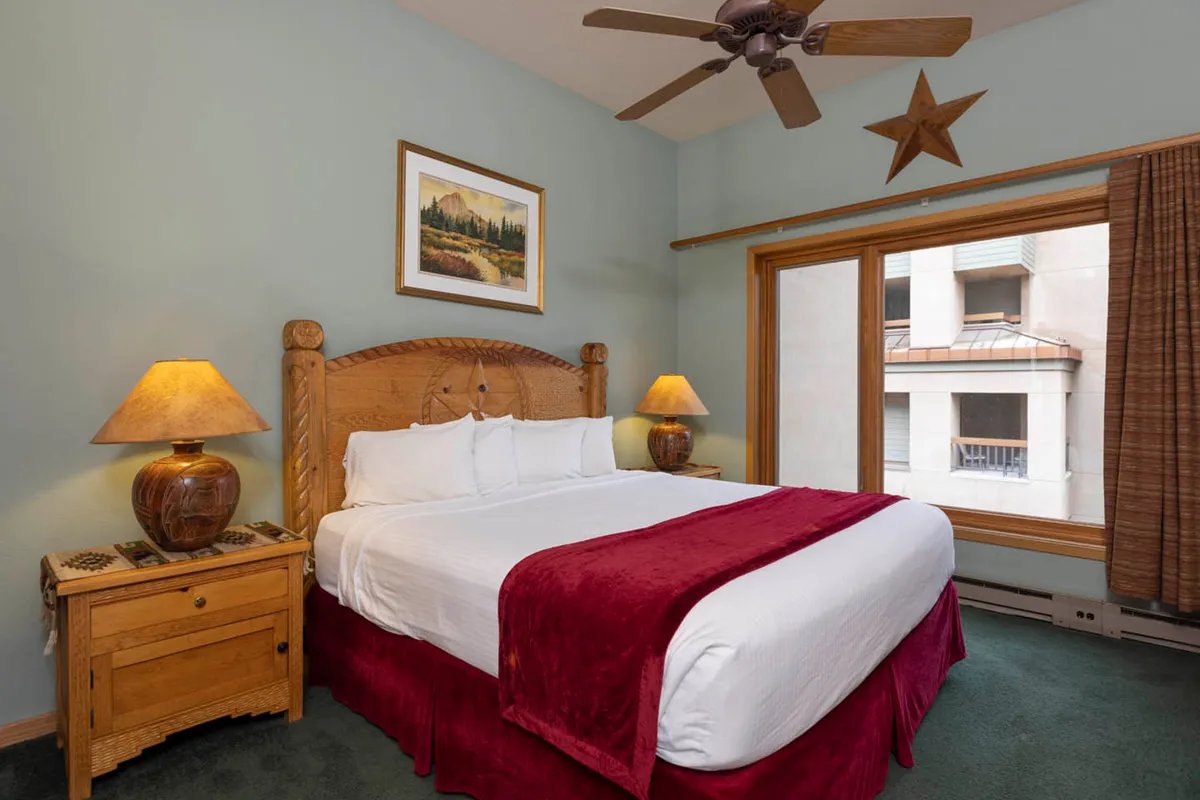 An inviting bedroom with a wooden headboard, plush bed topped with a red throw blanket, matching lamps on bedside tables, and a window with curtains letting in natural light. The space radiates comfort and warmth, with simple, rustic decor inspired by alpine surroundings.