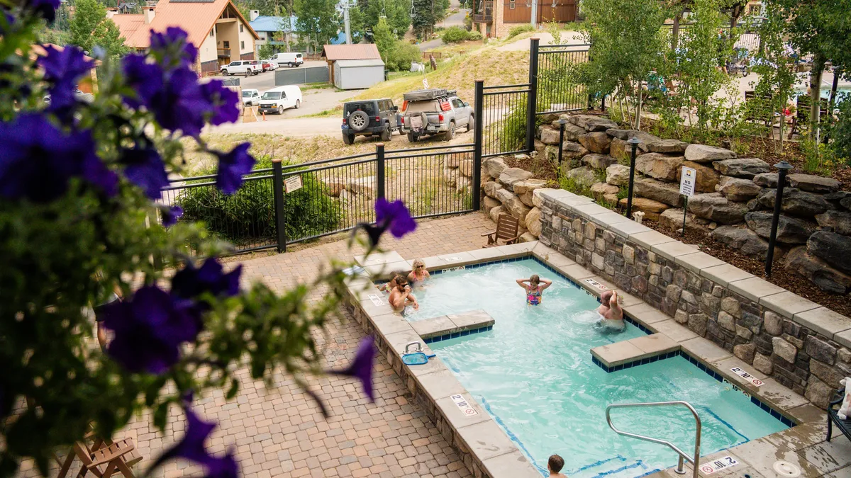 This exterior shot captures guests soaking in a rooftop hot tub, surrounded by lush flowers and ample poolside seating. In the background, the sun dips behind mountain peaks, bathing the scene in a warm, inviting light. The communal setting invites relaxation and socializing, perfectly illustrating the resort's signature amenity.
