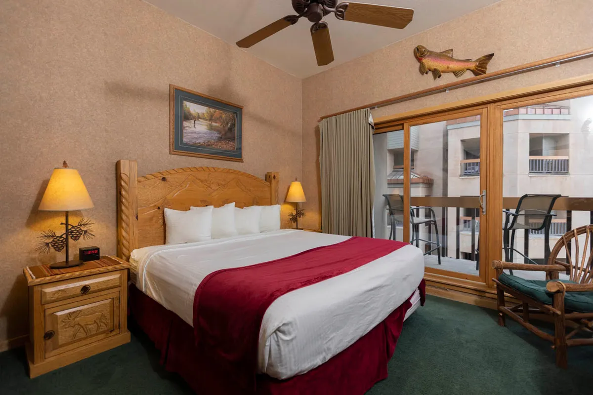 A cozy bedroom featuring a wooden bed frame with a red bedspread, a sliding glass door opening onto a private balcony, a wooden nightstand with a lamp, and a framed landscape painting above the bed. The space feels bright and welcoming, with natural light streaming in, and mountain-inspired decor touches.