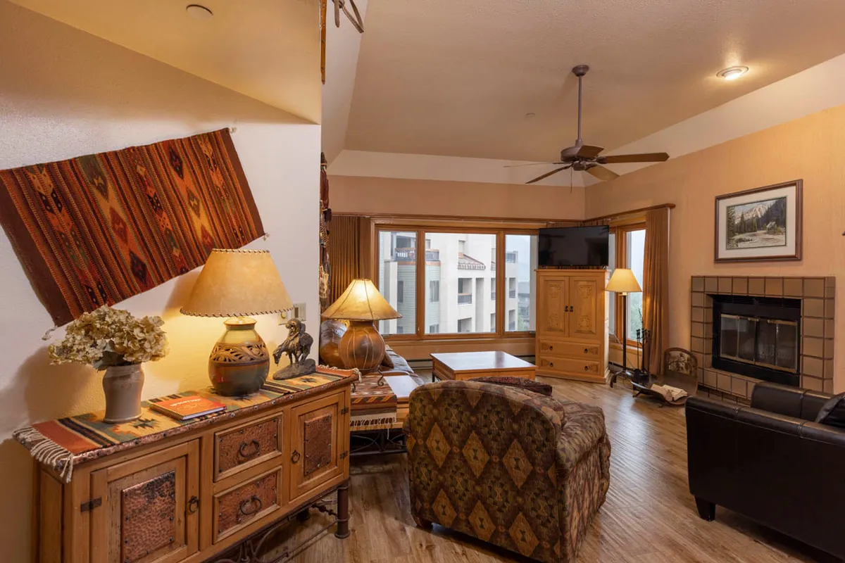 The image showcases a cozy, warmly lit condo living space featuring inviting wooden furniture, colorful decorative textiles, a roaring fireplace, and expansive windows that reveal striking mountain vistas. The scene suggests a comfortable spot for gathering, relaxing, and soaking in the natural beauty right from indoors.
