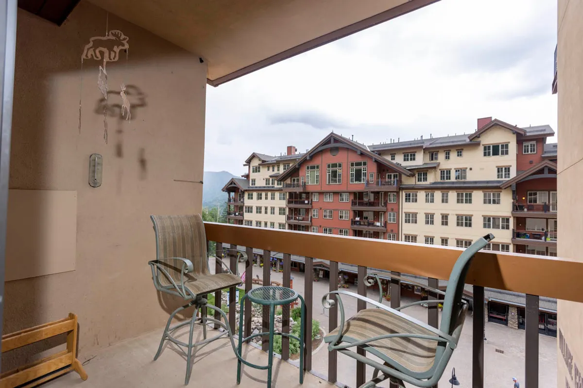 This exterior image captures a balcony space furnished with chairs, looking out onto surrounding buildings adorned with decorative wall art. The area is bathed in calm natural light, suggesting an inviting place to relax, people-watch, or enjoy a morning coffee while soaking in the atmosphere of the village center.
