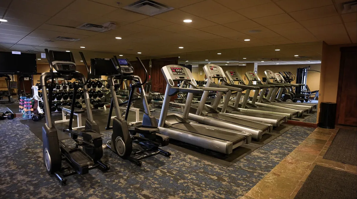An interior fitness center with elliptical machines, treadmills, a dumbbell rack, and expansive mirrors offers a bright, modern space for workouts. The equipment is neatly arranged, and the mood is energetic and motivating.