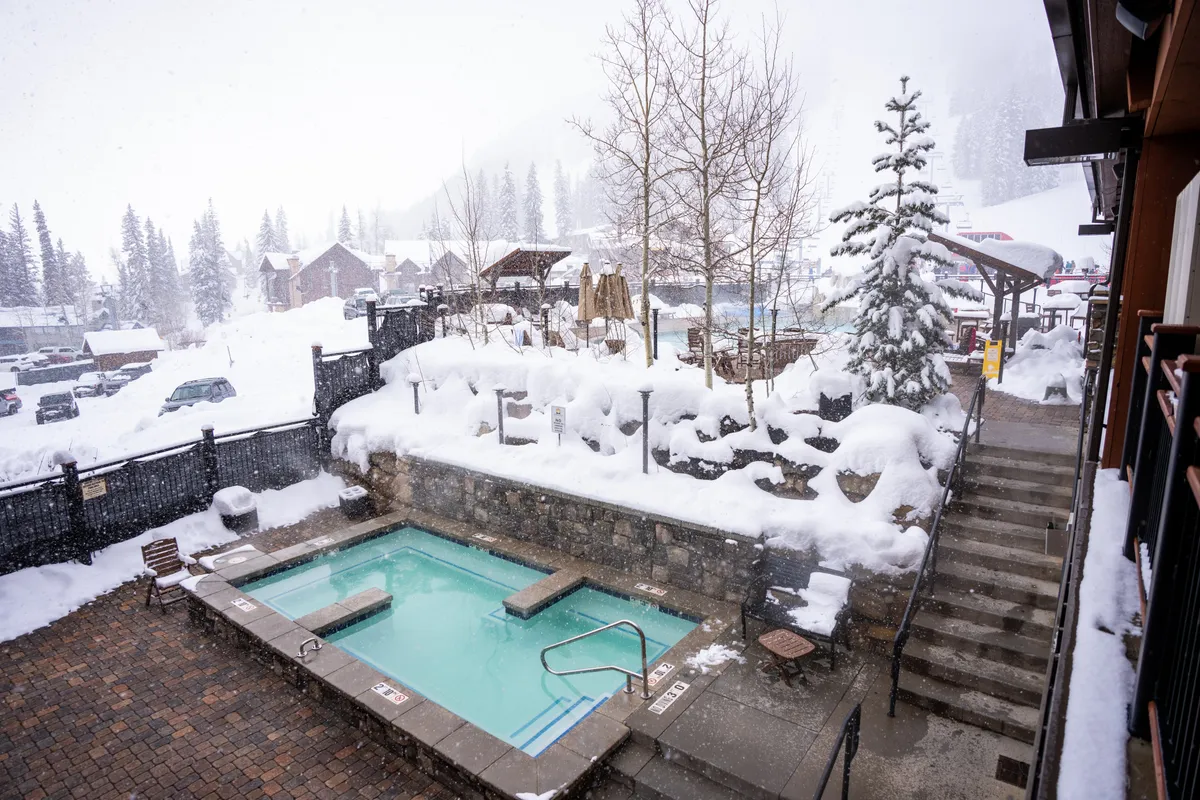 This exterior shot captures a snow-dusted hot tub surrounded by stone walls and steps, set amidst serene, snow-covered trees. The tranquil ambiance is enhanced by the wintry mountain setting, evoking the experience of soaking outdoors with panoramic alpine views.