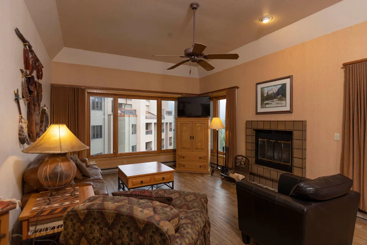 This image shows a spacious living area with large windows that allow natural light to pour in, offering stunning views of the surrounding mountains. The room features a cozy fireplace, a comfortable coffee table, and inviting sofas arranged for socializing and relaxation.