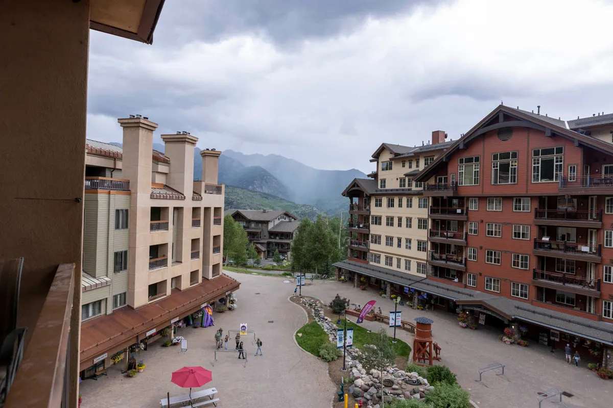 A lively exterior shot with people strolling through a vibrant plaza lined with buildings of diverse architecture, outdoor seating, and a pathway leading toward majestic mountains in the background. The mood is social and inviting, showcasing community activity against a scenic mountain backdrop.