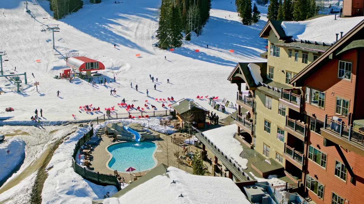 Featuring an outdoor pool and hot tub with adjacent ski slopes, this lively exterior scene blends the excitement of winter sports with the relaxation of resort amenities. People are seen skiing and socializing around the inviting pool area with mountain views all around.