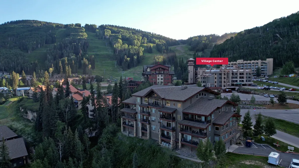 The lodge-style Village Center building with its prominent sign, surrounded by snow and framed by the majestic mountains. The parking area and nearby village hint at easy access and a welcoming, adventurous vibe.