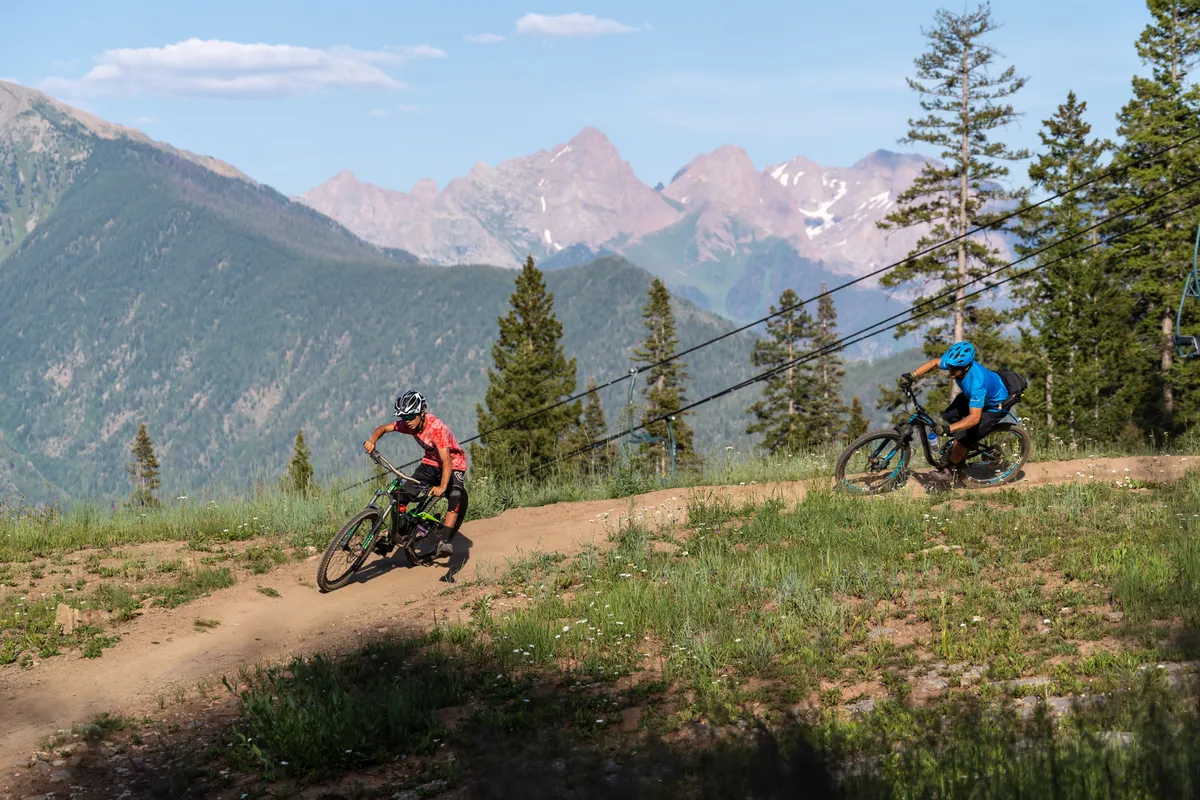 A dynamic outdoor scene featuring mountain bikers navigating a trail surrounded by lush pine trees and dramatic mountain landscapes under a clear blue sky. The image captures the thrill of mountain biking in the Rockies and hints at the wide range of outdoor activities available.